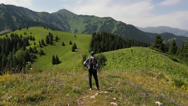 A drone flies picturesquely around a backpacker standing and admiring the stunning mountain views. A 360-degree view of a vast green valley with a spruce forest and mountains