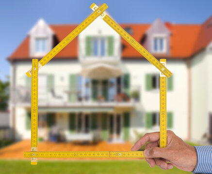 Ein Meterstab ist gefaltet in Form von einem Haus vor einem Neubau - FOTOMONTAGE