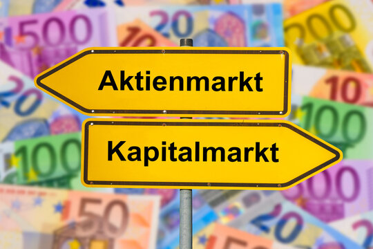Zwei Wegweiser &uuml;ber einer Menge Euro-Bargeld zeigen in unterschiedliche Richtungen und sind beschriftet mit Aktienmarkt und Kapitalmarkt - FOTOMONTAGE