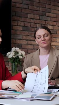 Two businesswomen review floor plan papers during office meeting, one in red blazer points and passes documents while seated at table with color swatches, coffee cup, brick wall backdrop.