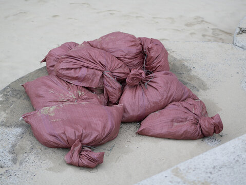 A pile of stacked purple-red woven plastic sacks filled with sand, tied at the ends and placed on a concrete surface near sand.