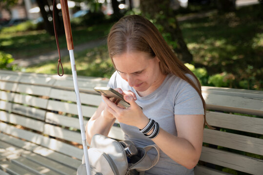 Blond haired visually impaired woman with white cane sitting outdoor and using a smartphone