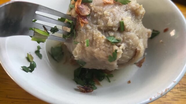 A large meatball being flipped over with a fork, revealing its texture beneath fried shallots and parsley.