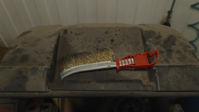 A wire scratch brush with an orange plastic handle lying on a dusty surface.