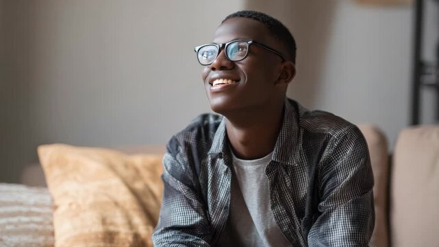 Young man smiles while wearing glasses. He sits relaxed on a couch indoors. Soft lighting highlights his joyful expression. Casual outfit adds to cozy, everyday feel. Background features muted tones