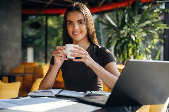 Young woman with long brown hair holding a white coffee cup while sitting at a table with a laptop and notebooks in a modern cafe with greenery in the background