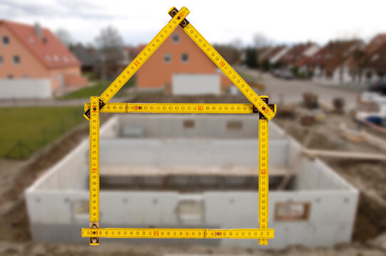 Ein Meterstab ist gefaltet in Form von einem Haus vor einem Neubau - FOTOMONTAGE