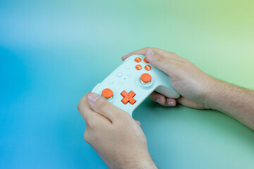 Hands holding a wireless mint green and orange game controller over a colorful blue and green gradient background, video game concept with copy space © Yuliia