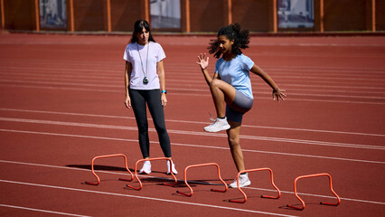 Fototapeta premium Teenage female athlete performing high knee drill over mini hurdles as adult female coach observes. Concept of hurdle coordination, youth fitness education, summer sport coaching.
