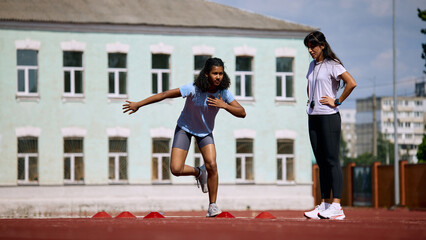 Fototapeta premium Teenage female athlete sprinting between cones as adult female coach observes. Concept of sprint acceleration, youth performance monitoring, summer sport competition.