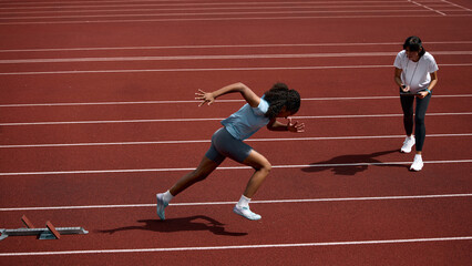 Naklejka na ściany i meble Black teenage female sprinter accelerating from starting blocks as adult female coach observes. Concept of sprint start acceleration, teenage performance development, summer sport coaching.
