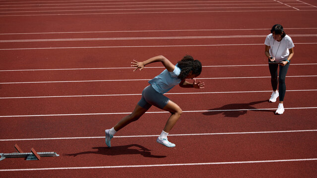 Black teenage female sprinter accelerating from starting blocks as adult female coach observes. Concept of sprint start acceleration, teenage performance development, summer sport coaching.