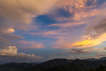 Obraz premium Golden pink sunset sky with moon above layered mountain ridge and soft clouds, peaceful natural landscape with warm light and pastel tones