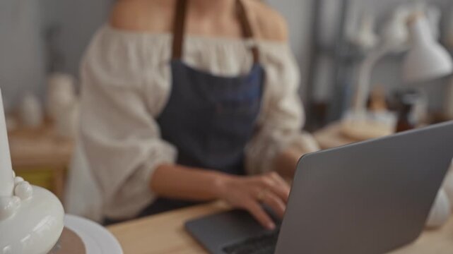 Woman in apron at pottery studio with laptop, hands typing on keyboard and off shoulder blouse visible; concentration.