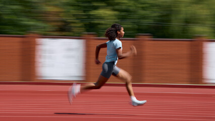 Obraz premium Panning shot of teenage female runner sprinting on outdoor athletics track with motion blur. Concept of youth athletic performance, running motion campaign, summer sport advertising.