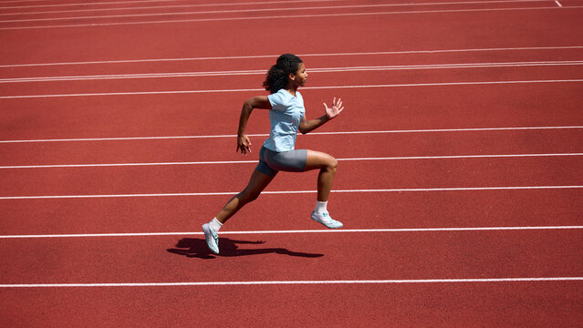 Black teenage female runner practicing sprint strides on outdoor athletics track. Concept of running technique, youth fitness development, summer sport training.