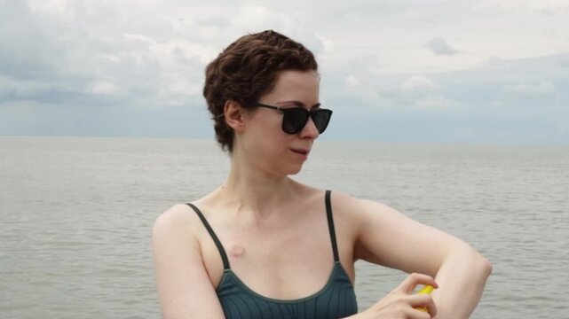 cancer survivor sprays sunscreen on her arm by the sea, showing sun protection, self care and safe summer travel during post treatment recovery and a calming beach escape