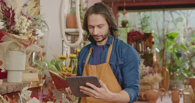 Flower shop owner uses a digital tablet while working among floral displays, highlighting inventory control, order management, entrepreneurship and efficient small business administration
