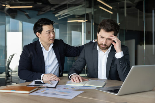 Two businessmen interacting in a modern office. One colleague empathetically offering comfort and support to another experiencing stress and anxiety while working at a desk with documents and a laptop