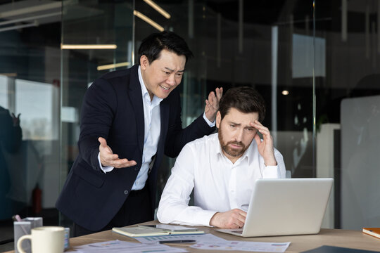 Experienced asian businessman expressing frustration and shouting at a frustrated caucasian employee working on a laptop. Illustrating workplace conflict. Harassment