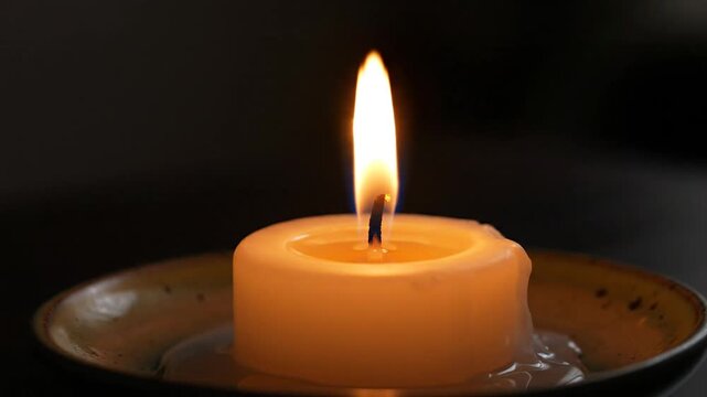 A close-up video of a single thick white candle burning on a small ceramic dish in a dark room. The flame flickers and dances gently, casting a warm glow.