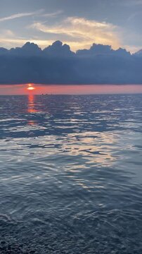 orange sun slowly sinks under a dramatic bank of storm clouds above gently rippling sea water, creating glowing reflections and a tranquil yet moody evening seascape