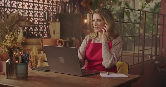 Flower shop owner speaks on her smartphone while using a laptop at her workspace, illustrating multitasking, business management, customer service and everyday retail operations
