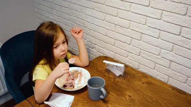 Young child eating breakfast at wooden dining table beside white brick wall in quiet home interior with warm light, gray mug, napkins, and simple morning setting