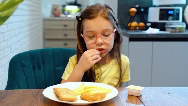 Child eating battered fish and thick-cut fries at home kitchen table, lifting food to mouth with visible bite action, seated beside plate, warm domestic meal scene with natural light