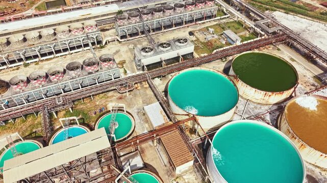 Aerial view shows large circular tanks filled with various liquids at an industrial site near a city. Pipes and equipment are visible around the tanks, indicating ongoing operations.