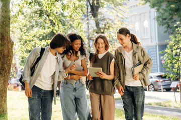 Fototapeta premium Group of four young adults, diverse friends, interacting with a tablet outdoors in a park, surrounded by trees and urban buildings in the background
