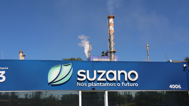 Fachada da unidade industrial da Suzano com logotipo e chamin&eacute; ao fundo, registrada em Suzano, S&atilde;o Paulo, Brasil, em 10 de agosto de 2026, sob c&eacute;u azul.