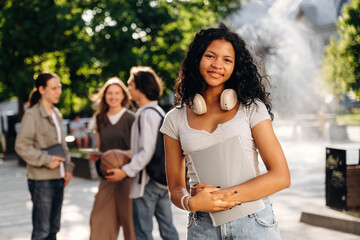 Obraz premium Young woman with curly hair wearing headphones holds a notebook while standing in a park with friends chatting and a fountain in the background