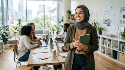Fototapeta premium A cheerful young woman wearing a hijab stands in a sunlit creative workspace holding a notebook while her diverse professional team collaborates on new business projects at a large wooden table
