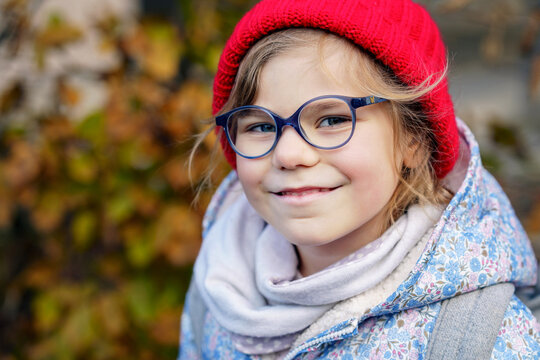 Portrait of a cute preschool girl with eye glasses and red hat outdoors. Happy funny child wearing new blue glasses. Autumn day in the city.