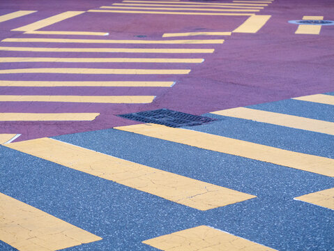 Close-up of a paved urban road intersection with yellow pedestrian crosswalk stripes, grey asphalt, and a reddish-purple surface texture.