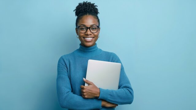 Bright smile radiates from a young black woman. She holds a laptop close, dressed in a soft blue sweater. Her glasses and natural curls add a warm, approachable charm
