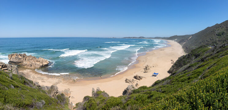 High resolution stitched panorama of Brenton on Sea beach, South Africa, featuring a wide angle panoramic view of turquoise ocean waves, a vast sandy shoreline, and green coastal fynbos hills.