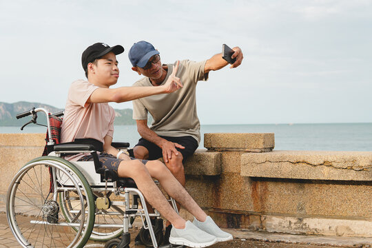 A happy young disabled man in a wheelchair posing with a V-sign gesture while taking a selfie with his caring male caregiver on smartphone near the beach, enjoying accessible travel lifestyle.