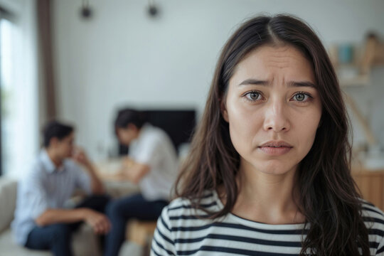 Young woman shows distress, eyes wide, brow furrowed. Two men sit in background, heads down, avoiding eye contact. Room feels tense, quiet, emotionally heavy. Use for stories of family conflict