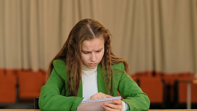 A focused schoolgirl checks a notebook page at a classroom desk, calm tones and natural light emphasizing emotion. It relates to study routine, concentration fatigue, and hidden depression.