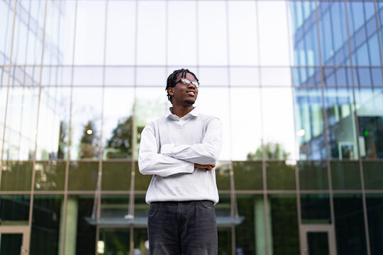 A young African American man with dreadlocks and glasses stands with his arms crossed in front of a modern glass building