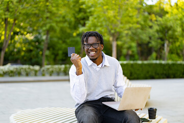 Fototapeta premium A smiling african american man sits outdoors with a laptop and credit card, ready for online transactions