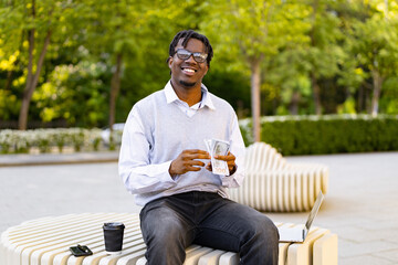 Obraz premium A smiling african american man in glasses sits on a modern bench outdoors with a laptop and coffee, counting US dollar bills
