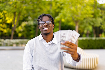 Fototapeta premium A smiling young African American man in glasses holds a fan of US dollar bills in his hand while standing outside in a park