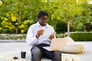 Obraz premium A smiling African American man sits outdoors on a modern bench, holding US dollar bills and looking at his laptop screen