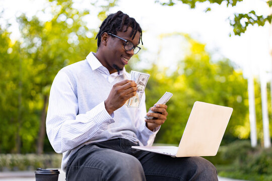 A smiling African American man sits outdoors with a laptop and counts a stack of US dollar bills, possibly managing finances or celebrating a success
