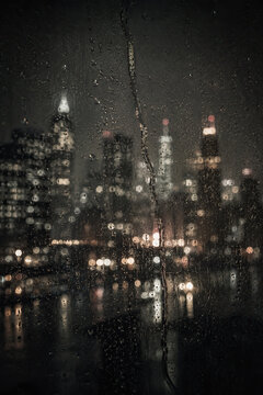 A captivating nighttime cityscape reflected in a textured glass window, showcasing a monochromatic urban scene with illuminated buildings and scattered water droplets.