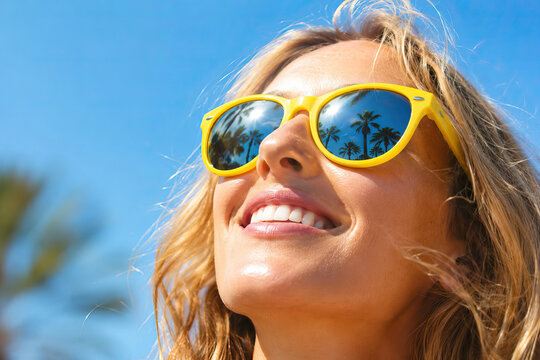 Close-up portrait of a person with blonde hair, wearing yellow sunglasses reflecting palm trees against a clear blue sky, smiling with a moment of happiness.