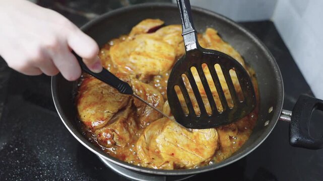 Close-up of marinated pork steaks cooking in savory sauce in a frying pan on a gas stove. A person uses a black spatula to flip the meat. Home cooking and delicious dinner food preparation.